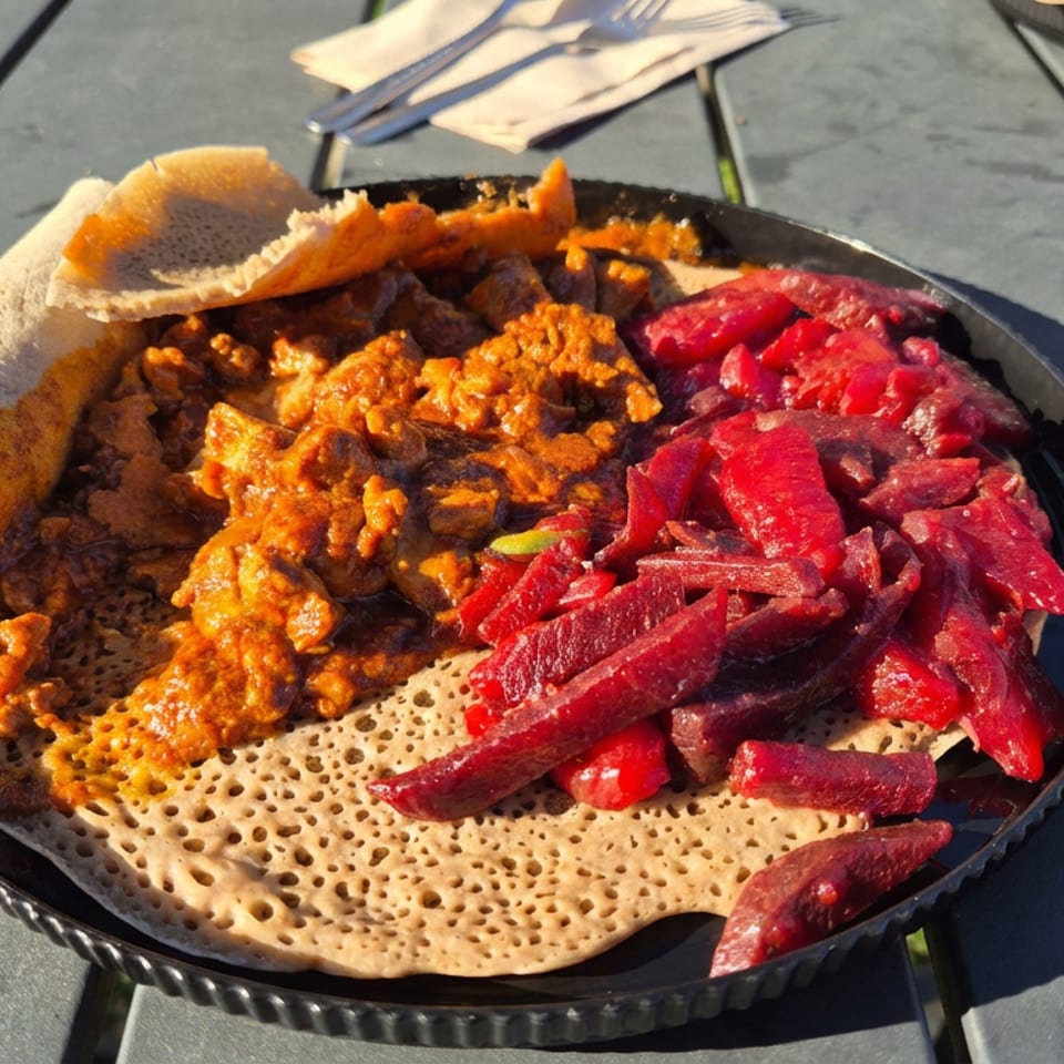 Ethiopian injera topped with spicy stew and bright red beet salad on a black plate outdoors.